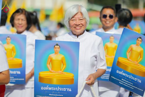หญิงสูงวัยผมสีขาวสวมชุดขาวถือป้ายภาพพระพุทธรูปสีทองในพิธีทางศาสนา
