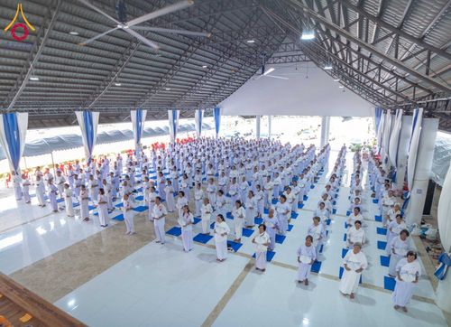 กลุ่มคนสวมชุดสีขาวจำนวนมากยืนเรียงแถวปฏิบัติธรรมภายในอาคาร