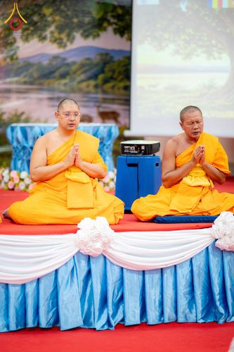 พระสงฆ์สองรูปนั่งพนมมือบนเวทีที่มีการตกแต่งด้วยผ้าสีฟ้าและขาว