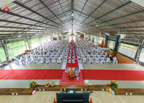 กลุ่มอุบาสกและอุบาสิกาสวมชุดขาวนั่งปฏิบัติธรรมภายในอาคารกว้างขวาง โดยมีพระภิกษุนั่งนำกิจกรรมอยู่ด้านหน้า