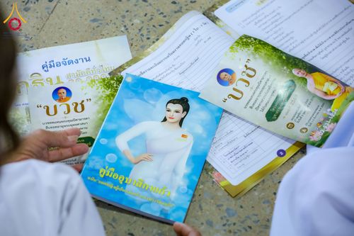 มือของผู้เข้าร่วมโครงการถือคู่มืออุบาสิกาแก้วและแผ่นพับประชาสัมพันธ์โครงการทอดกฐินสามัคคีวางอยู่บนพื้น