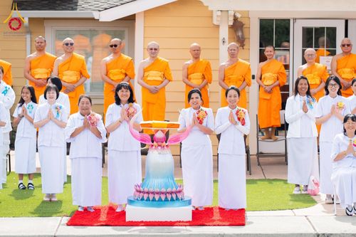 พระภิกษุสงฆ์และกลุ่มสาธุชนชุดขาวกำลังประกอบพิธีทางศาสนาพุทธ โดยมีพานพุ่มผ้าไตรตั้งอยู่ด้านหน้า