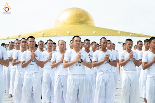 กลุ่มนักเรียนนายสิบตำรวจในชุดสีขาวพนมมือปฏิบัติธรรมกลางแจ้ง โดยมีอาคารทรงโดมสีทองเป็นฉากหลัง