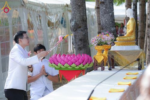 ชายสองคนสวมชุดสีขาวกำลังจุดเทียนบูชาพระพุทธรูปสีทองบนโต๊ะที่มีพานดอกไม้ประดับ