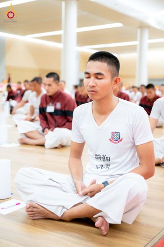 นักเรียนนายสิบตำรวจชายสวมเสื้อยืดสีขาวนั่งขัดสมาธิหลับตาทำสมาธิบนพื้นไม้ภายในห้องโถง