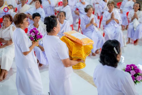 กลุ่มผู้หญิงในชุดสีขาวกำลังถือพานพุ่มผ้าไตรและช่อดอกไม้ในพิธีทางศาสนา