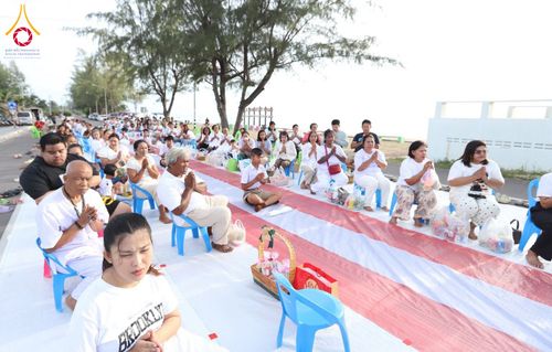 กลุ่มประชาชนสวมชุดสีขาวนั่งพนมมือเรียงแถวบนเสื่อริมชายหาดเพื่อรอตักบาตร