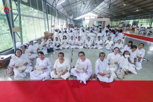 กลุ่มอุบาสิกาแก้วสวมชุดขาวนั่งเรียงแถวถือเอกสารภายในอาคารปฏิบัติธรรม