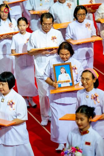กลุ่มคนสวมชุดสีขาวถือกรอบรูปและถาดเดินในขบวนพิธีการบนพรมสีแดง