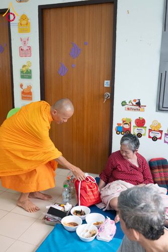 พระภิกษุส่งมอบถุงยังชีพให้หญิงสูงวัยภายในห้องที่มีการตกแต่งด้วยรูปภาพการ์ตูน