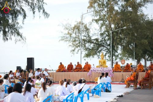กลุ่มคนสวมชุดขาวนั่งร่วมพิธีตักบาตรพระสงฆ์บนเวทีริมชายหาด