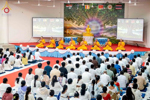 พระภิกษุสงฆ์และพุทธศาสนิกชนร่วมกิจกรรมทางศาสนาภายในหอประชุมที่มีการตกแต่งด้วยภาพพระพุทธรูปและธงชาติ