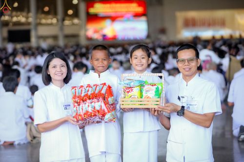 กลุ่มคนสี่คนถือของตักบาตรในชุดสีขาวภายในอาคารที่มีผู้คนมาร่วมพิธีจำนวนมาก