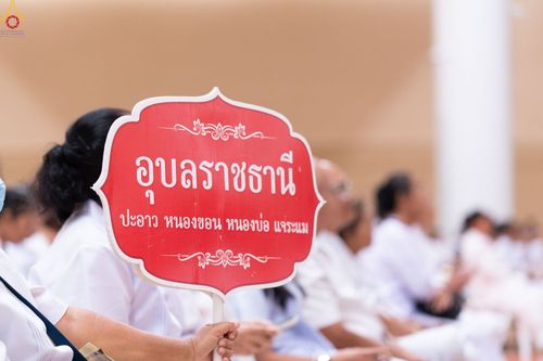 ป้ายสีแดงระบุข้อความอุบลราชธานีและชื่อพื้นที่ย่อย ถือโดยผู้เข้าร่วมงานในชุดสีขาวภายในอาคาร