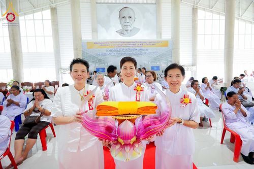 กลุ่มสตรีในชุดขาวถือพานผ้าไตรในพิธีทางศาสนาภายในอาคารที่มีผู้เข้าร่วมงานนั่งอยู่โดยรอบ