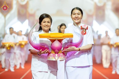 หญิงสองท่านถือพานพุ่มดอกบัวสีชมพูวางผ้าไตรจีวรสีเหลืองทองในพิธีทางศาสนา