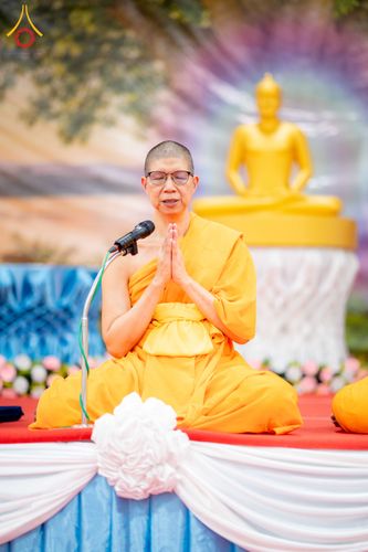 ภิกษุณีสวมแว่นตานั่งพนมมือสวดมนต์หน้าไมโครโฟนบนเวทีที่มีพระพุทธรูปเป็นฉากหลัง