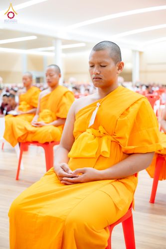 พระภิกษุสงฆ์ห่มจีวรสีเหลืองนั่งหลับตาทำสมาธิบนเก้าอี้พลาสติกสีแดงภายในห้องโถง