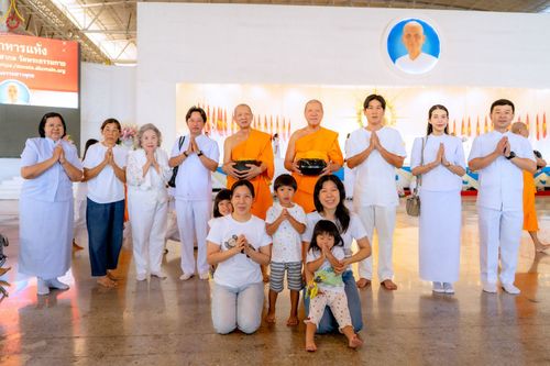 กลุ่มคนและพระภิกษุสงฆ์ยืนพนมมือร่วมพิธีตักบาตรภายในอาคาร