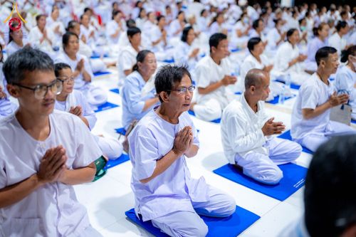 กลุ่มคนสวมชุดสีขาวนั่งพนมมือปฏิบัติธรรมบนเบาะรองนั่งสีน้ำเงินในพื้นที่อบรม