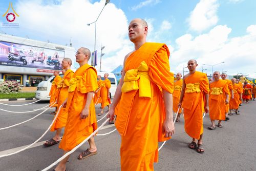 คณะสงฆ์หลายรูปสวมจีวรสีส้มเดินเรียงแถวบนถนนและถือสายสิญจน์ในมือ