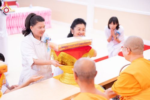 หญิงไทยในชุดสีขาวกำลังประเคนผ้าไตรบนพานพุ่มสีทองแด่พระภิกษุสงฆ์ในพิธีทางศาสนา