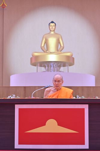 พระภิกษุสงฆ์กำลังแสดงธรรมหน้าไมโครโฟนโดยมีพระพุทธรูปสีทองเป็นฉากหลัง