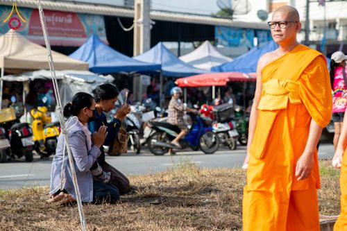 พุทธศาสนิกชนนั่งพนมมือไหว้พระภิกษุที่กำลังเดินผ่านบริเวณงานกลางแจ้ง
