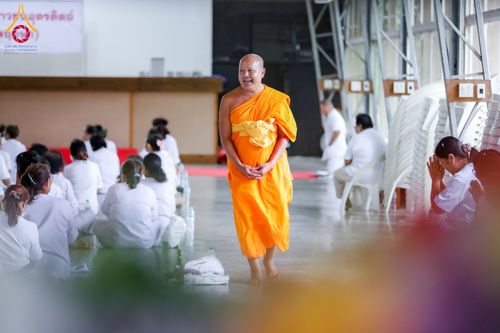 พระภิกษุเดินยิ้มแย้มในอาคารที่มีกลุ่มคนชุดขาวนั่งอยู่