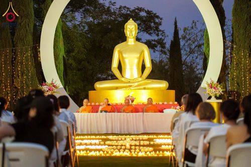 พระภิกษุสงฆ์นั่งบนเวทีหน้าพระพุทธรูปทองคำและกลุ่มผู้เข้าร่วมกิจกรรมนั่งสมาธิท่ามกลางแสงเทียนในยามเย็น