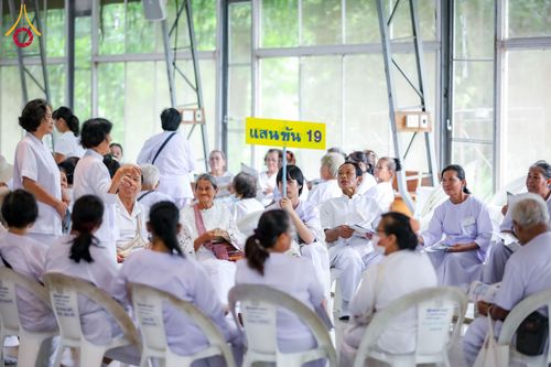 กลุ่มคนสวมชุดสีขาวนั่งทำกิจกรรมร่วมกันภายในอาคาร มีป้ายสีเหลืองเขียนว่า แสนขัน 19