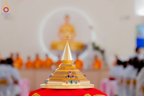 เจดีย์จำลองสีทองประดับอัญมณีวางอยู่บนพานสีแดง ด้านหลังเป็นภาพเบลอของพระพุทธรูปและพระภิกษุสงฆ์