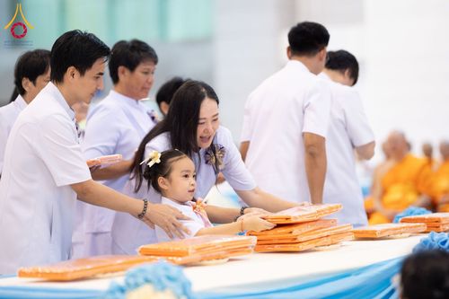 กลุ่มคนสวมชุดสีขาวกำลังวางผ้าไตรจีวรสีส้มบนโต๊ะยาวในพิธีทางศาสนา