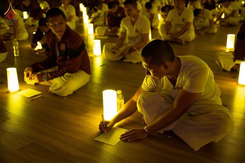 กลุ่มคนสวมชุดสีขาวนั่งปฏิบัติธรรมบนพื้นไม้ท่ามกลางแสงไฟจากโคมไฟทรงกระบอกในห้องโถง