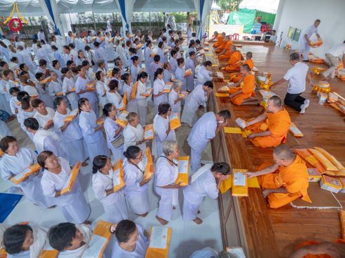 กลุ่มอุบาสิกาชุดขาวกำลังเข้าแถวถวายสังฆทานแด่พระภิกษุสงฆ์ที่นั่งอยู่บนอาสนะไม้