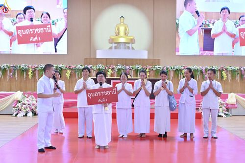 กลุ่มคนสวมชุดสีขาวบนเวทีงานสัมมนา มีผู้ถือป้าย อุบลราชธานี คันที่ 1 และมีผู้ชายพูดผ่านไมโครโฟน