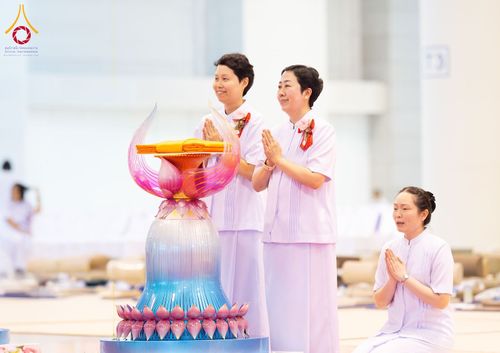อุบาสิกาสวมชุดสีขาวพนมมือในพิธีทางศาสนาหน้าพานพุ่มดอกบัวที่มีผ้าไตรวางอยู่