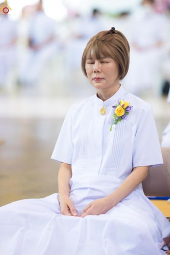 หญิงสาวสวมชุดขาวนั่งหลับตาทำสมาธิ ติดเข็มกลัดดอกไม้ที่หน้าอก