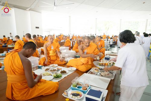 พระภิกษุสงฆ์กำลังฉันภัตตาหารภายในหอฉัน โดยมีอาสาสมัครชุดขาวช่วยจัดเตรียมอาหาร