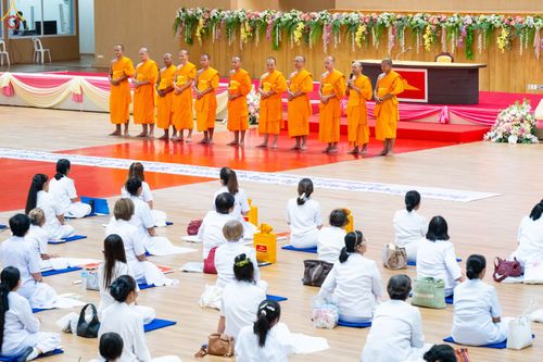 พระภิกษุสงฆ์ยืนบนเวทีและสาธุชนชุดขาวนั่งปฏิบัติธรรมในหอประชุม