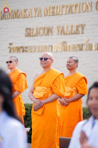 พระภิกษุสงฆ์สามรูปสวมจีวรสีส้มยืนอยู่หน้าป้ายชื่อวัดพระธรรมกายซิลิคอนวัลเลย์