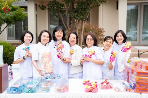กลุ่มผู้หญิง 7 คนสวมชุดสีขาวถือดอกไม้ประดิษฐ์ยืนยิ้มหน้าโต๊ะที่มีของตกแต่งและกล่องของขวัญ