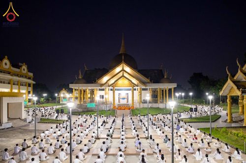 กลุ่มคนสวมชุดสีขาวนั่งสมาธิบนลานกว้างหน้าอาคารทรงไทยในยามค่ำคืน