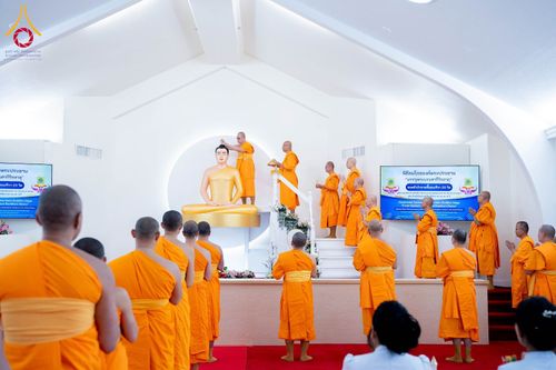 พระภิกษุสงฆ์ในจีวรสีส้มกำลังประกอบพิธีบนแท่นประดิษฐานพระประธานสีทองภายในห้องโถงสีขาวที่มีผู้เข้าร่วมพิธีจำนวนมาก