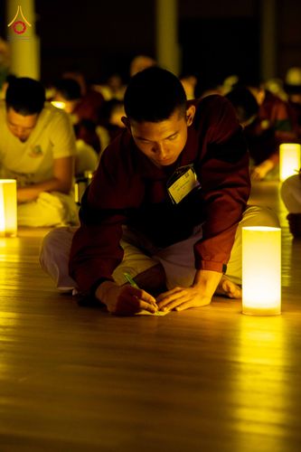 ชายหนุ่มสวมชุดปฏิบัติธรรมนั่งเขียนหนังสือบนพื้นไม้ท่ามกลางแสงไฟจากโคมไฟเทียนในห้องโถง