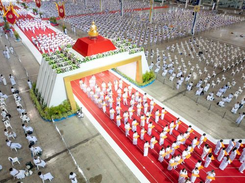 กลุ่มคนในชุดขาวเดินแถวบนพรมแดงเข้าสู่พื้นที่พิธีภายในอาคารขนาดใหญ่ที่มีผู้คนนั่งประจำที่จำนวนมาก