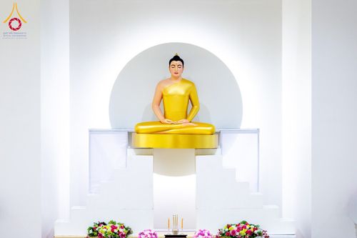พระพุทธรูปสีทองปางสมาธิประดิษฐานบนฐานสีขาว มีฉากหลังเป็นวงกลมสีขาวและประดับด้วยดอกไม้สด