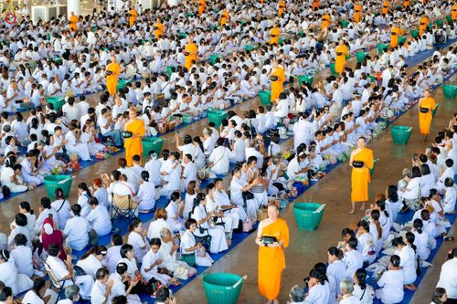 พุทธศาสนิกชนชุดขาวจำนวนมากนั่งเรียงแถวรอตักบาตรพระภิกษุในอาคารกว้างขวาง