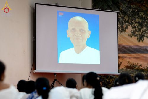 ภาพฉายบนจอโปรเจกเตอร์แสดงภาพบุคคลโกนศีรษะสวมชุดขาว โดยมีกลุ่มคนนั่งหันหลังชมอยู่ด้านหน้า