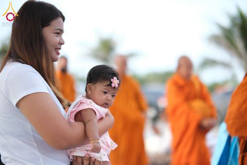 หญิงสาวอุ้มทารกน้อยในอ้อมแขนขณะร่วมพิธีตักบาตรที่มีพระสงฆ์เดินผ่าน